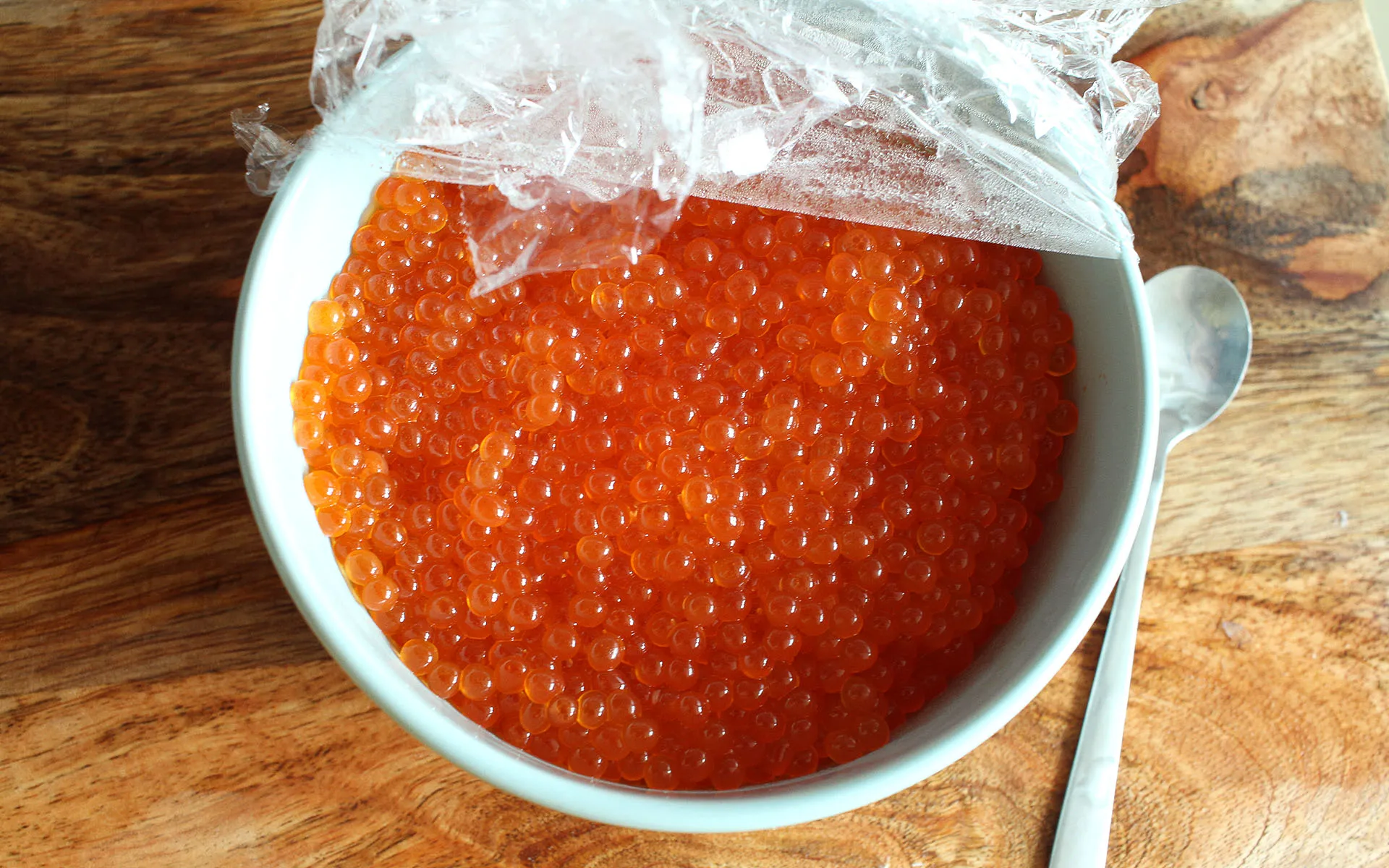 Vivid red salmon roe eggs in a stainless bowl on a wooden benchtop.