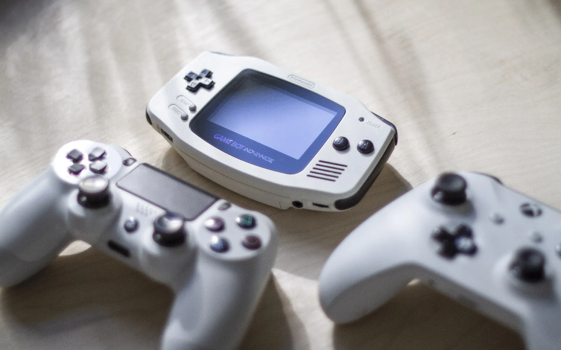 White Gameboy Advance next to a white Xbox one and PS4 controllers.